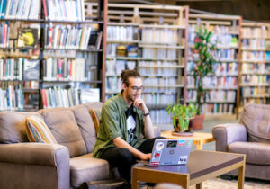 男大学生坐在图书馆的沙发上，拿着笔记本电脑